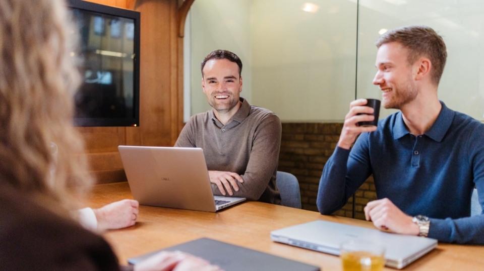 Drie collega’s zitten aan een vergadertafel met laptops en koffie en lachen tijdens een informeel overleg.