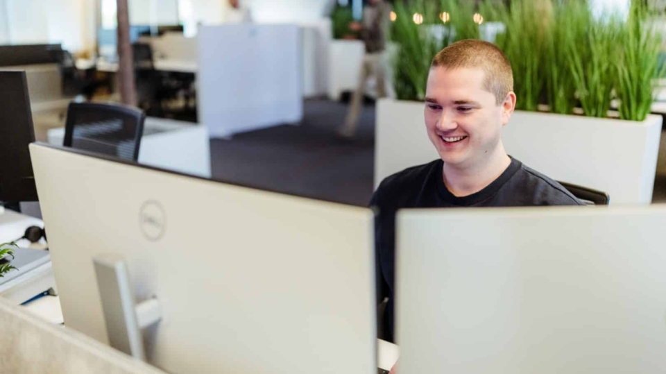 Een man glimlacht terwijl hij achter zijn bureau zit in een kantooromgeving, met planten op de achtergrond.