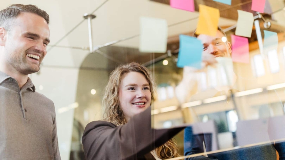 Drie collega’s plakken gekleurde post-its op een glazen wand tijdens een brainstorm en lachen samen.