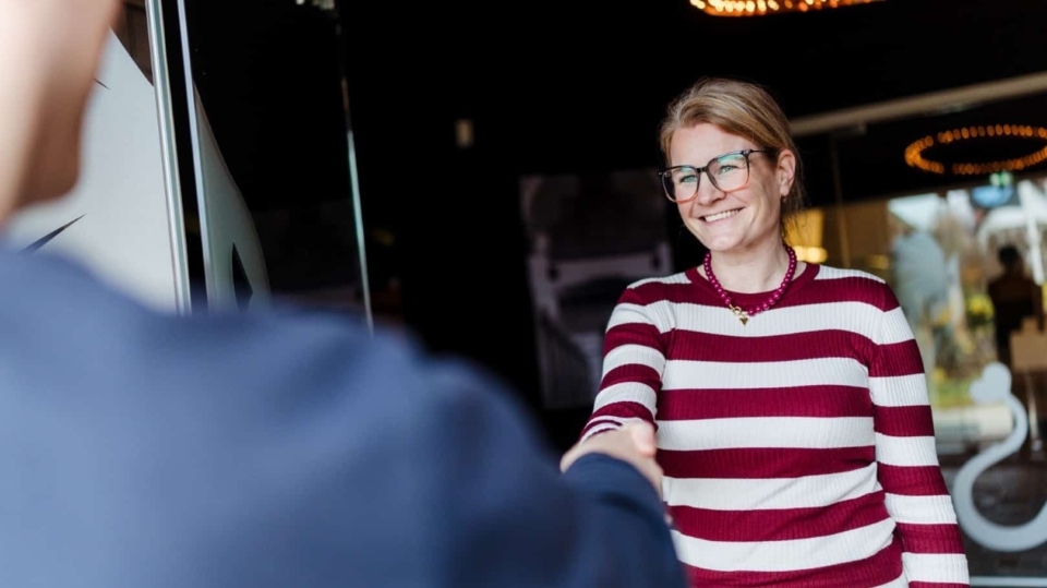 Een vrouw schudt glimlachend de hand van een bezoeker bij de ingang van een kantoor.