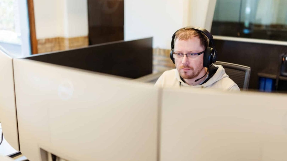 Een man met headset werkt geconcentreerd achter twee beeldschermen in een kantooromgeving.