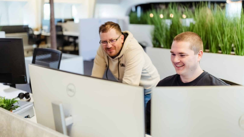 Twee collega’s werken samen achter computers in een kantoor en glimlachen terwijl ze naar het scherm kijken.