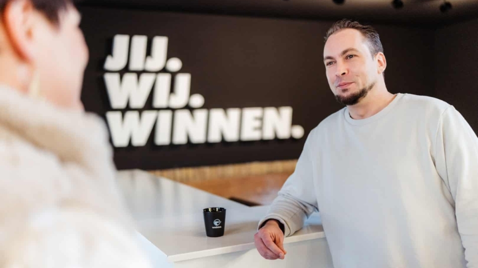 Een man staat bij een balie in een kantoorruimte en voert een gesprek met een bezoeker, met de tekst ‘Jij. Wij. Winnen.’ zichtbaar op de achtergrond.