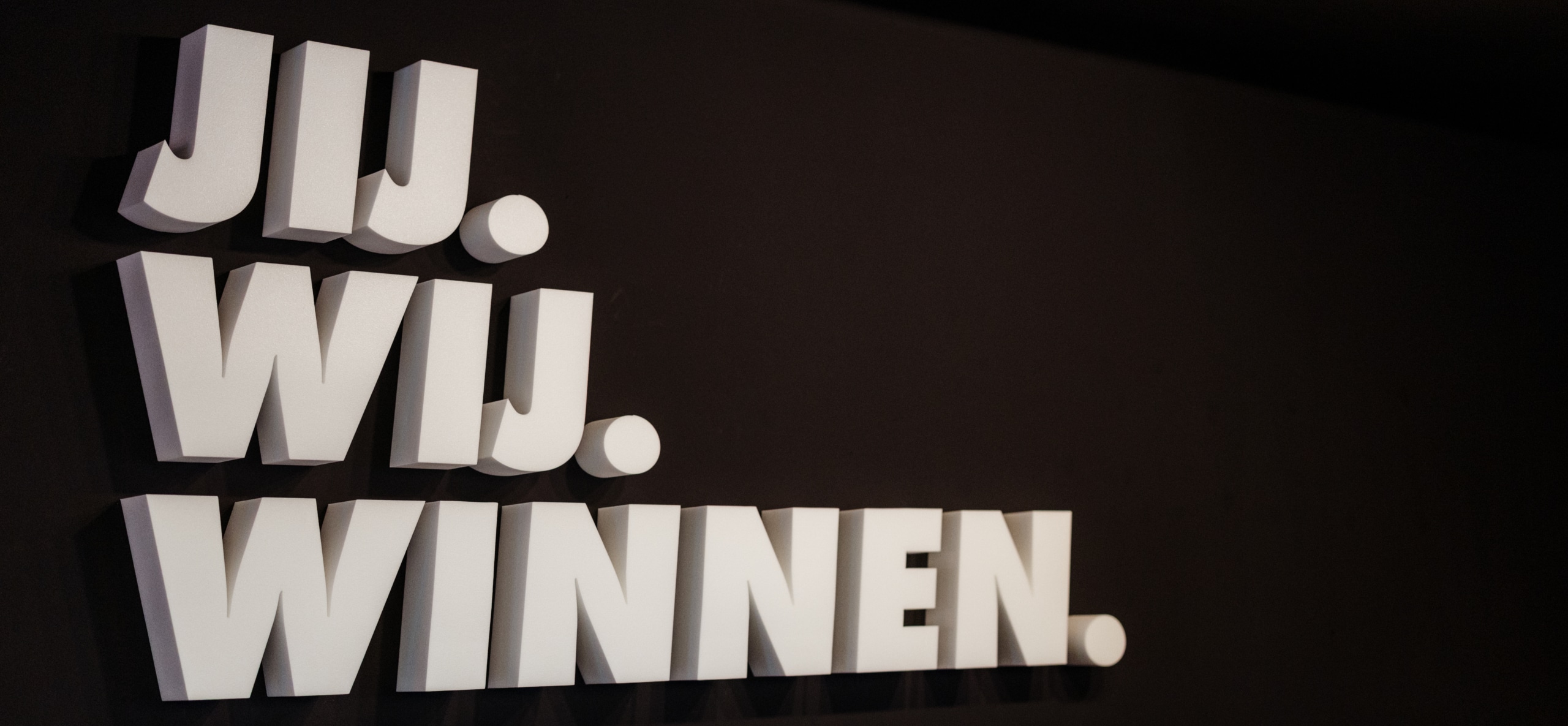 Tekst ‘Jij. Wij. Winnen.’ op een donkere muur als slogan of bedrijfsuiting in een kantoorruimte.