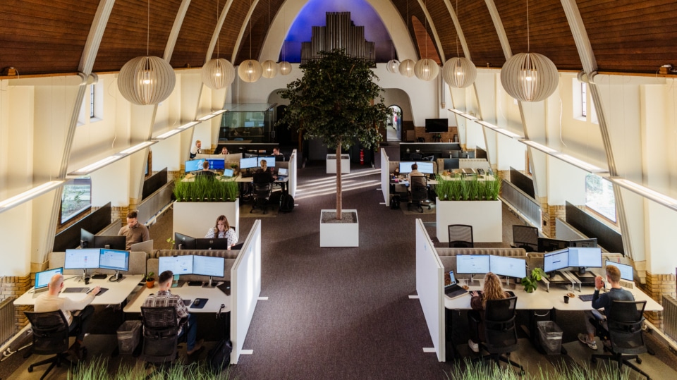 Een breed uitzicht op de open kantoorruimte van Webton in Hengelo. Met meerdere bureaus en medewerkers aan hun werkstations. In het midden staat een grote plant en het plafond heeft een hoge, houten structuur. De ruimte is goed verlicht en ziet er modern en ruim uit.