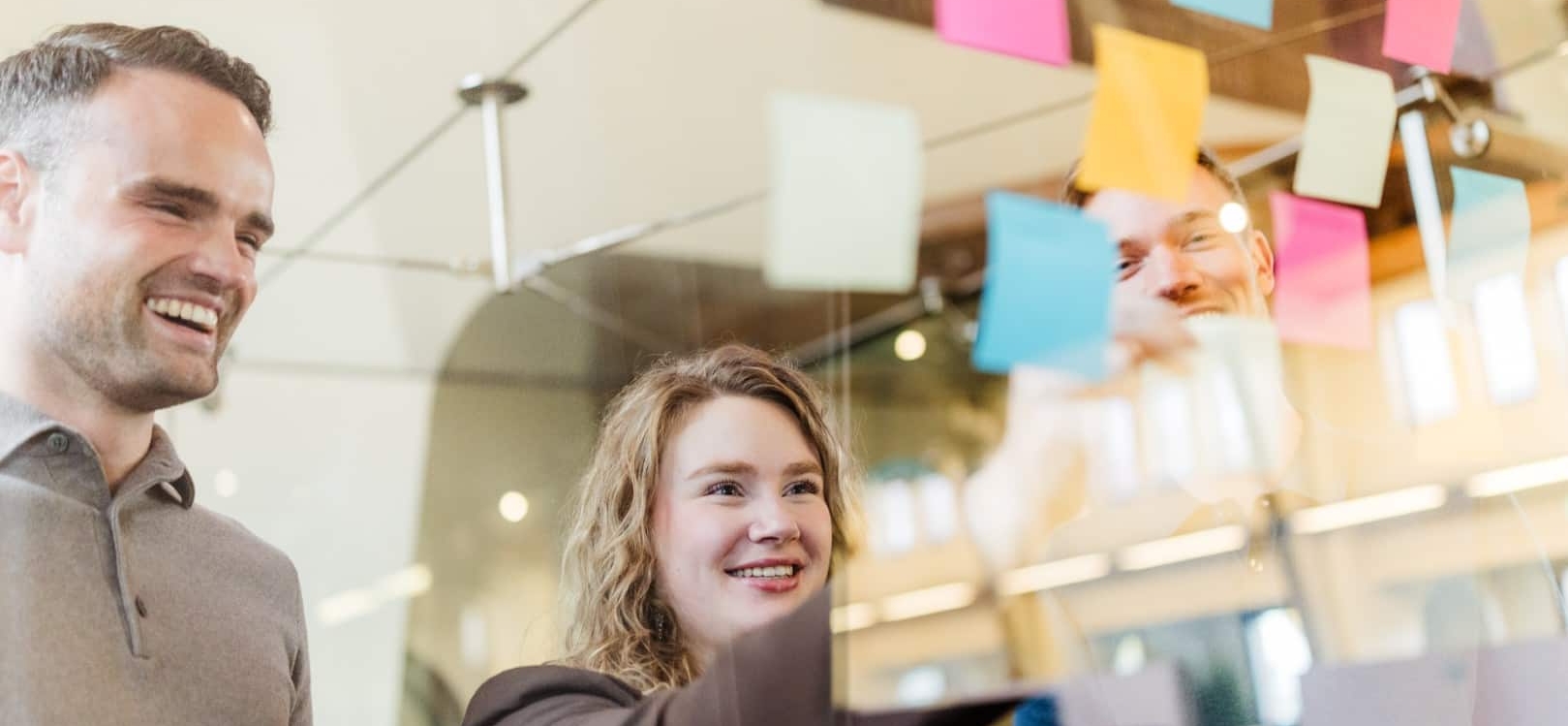 Drie collega’s plakken gekleurde post-its op een glazen wand tijdens een brainstorm in een kantooromgeving.