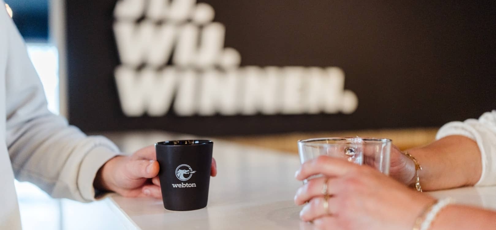 Twee personen staan bij een balie met een kopje koffie en een glas water, met de tekst ‘Jij. Wij. Winnen.’ op de achtergrond.