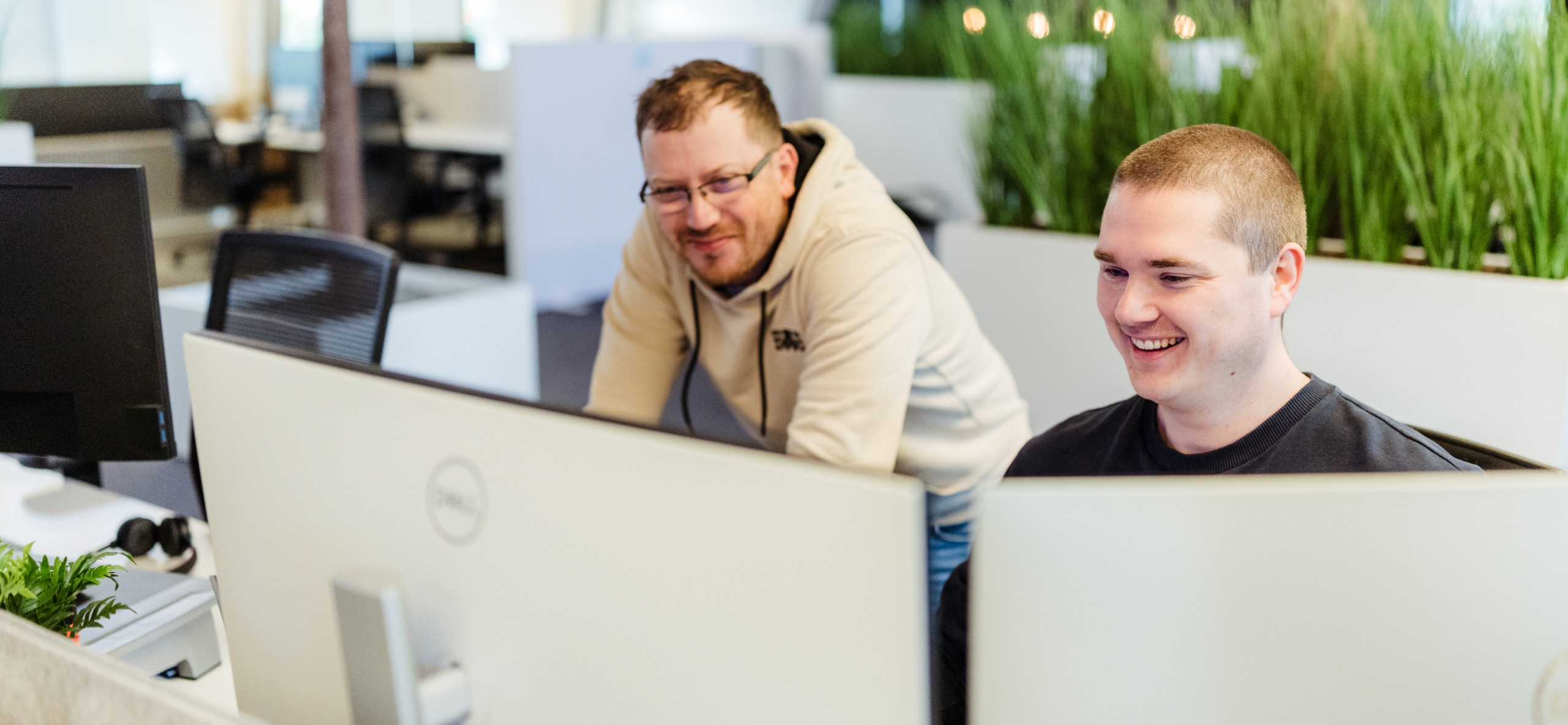 Twee collega’s werken samen achter computerschermen in een moderne kantooromgeving, waarbij één over de schouder meekijkt.