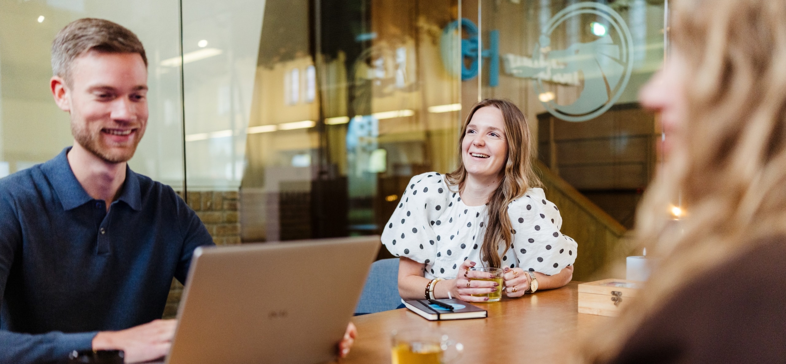 Drie collega’s zitten rond een tafel met een laptop tijdens een overleg. Ze lachen en lijken een informeel gesprek te voeren.