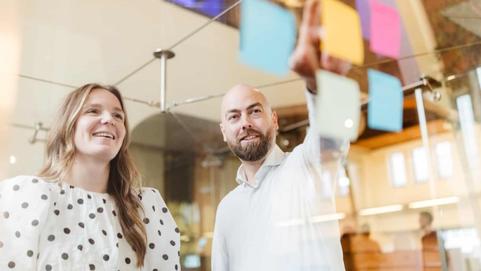 Een vrouw en een man staan in een vergaderruimte en kijken naar een reeks kleurrijke post-its die op een glazen wand zijn geplakt. De vrouw glimlacht terwijl de man wijst naar een post-it.