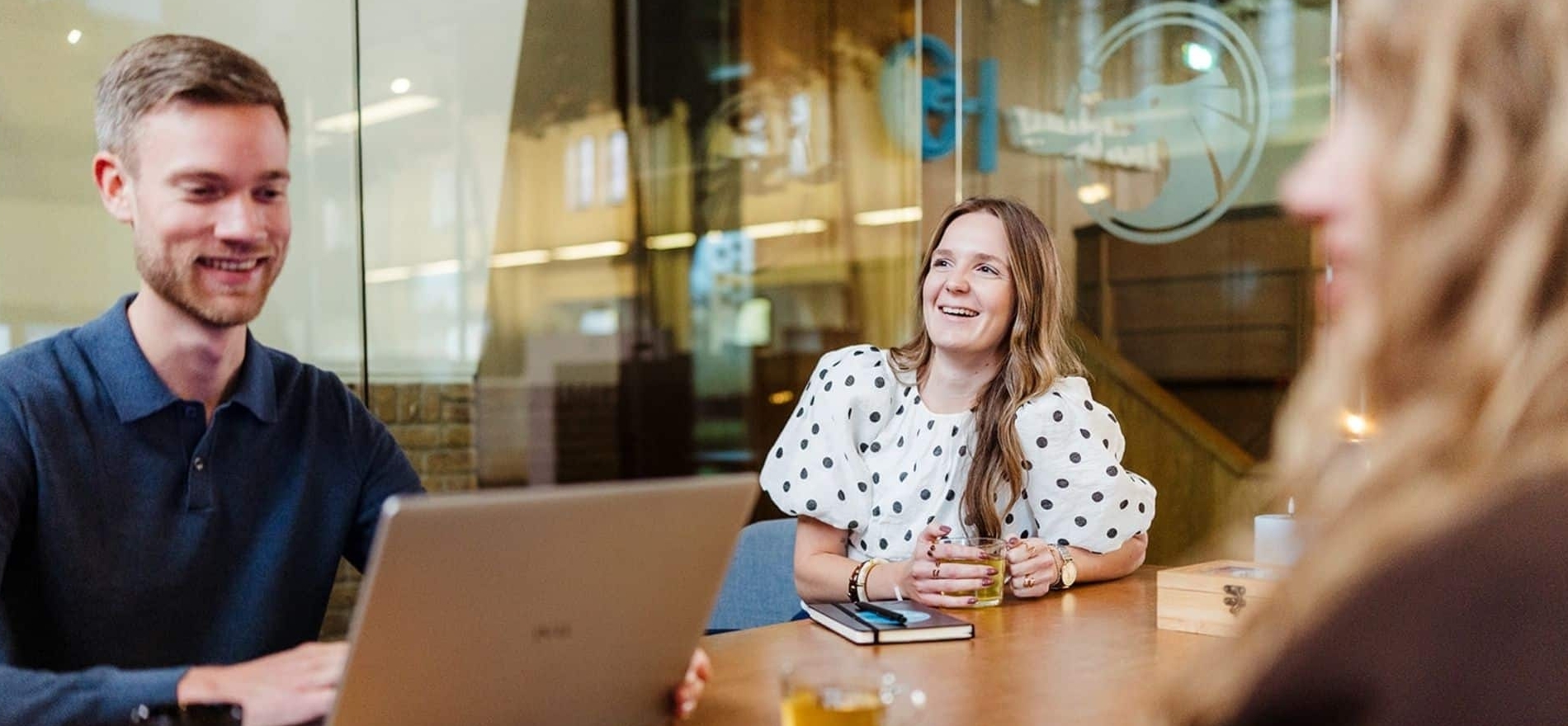 Drie collega’s zitten rond een tafel met een laptop tijdens een overleg. Ze lachen en lijken een informeel gesprek te voeren.