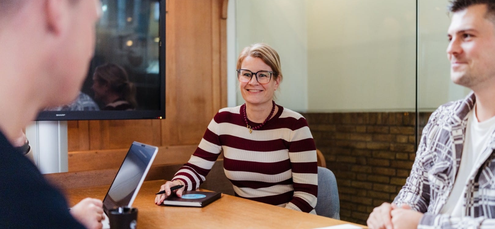 Een vrouw in een gestreept shirt glimlacht terwijl ze praat met collega's, waarbij ze in een vergaderruimte staan. Ze lijkt te genieten van het gesprek.