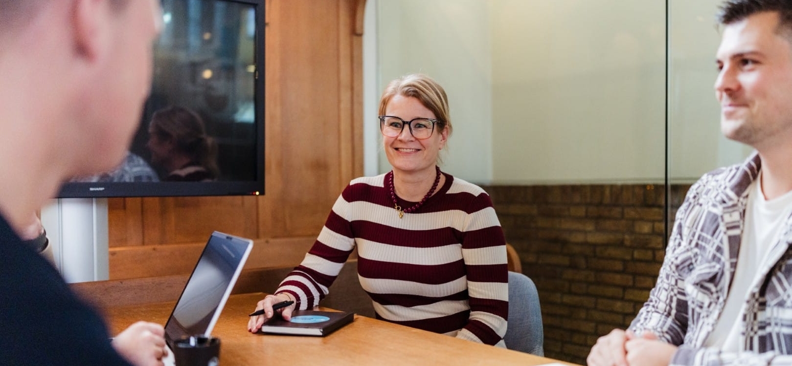 Een vrouw in een gestreept shirt glimlacht terwijl ze praat met collega's, waarbij ze in een vergaderruimte staan. Ze lijkt te genieten van het gesprek.
