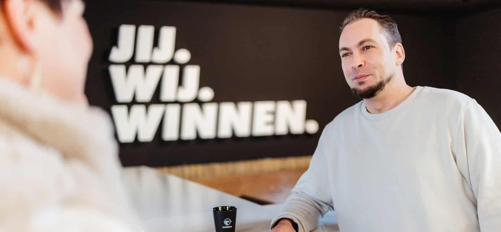 Man in gesprek bij een balie met op de achtergrond de tekst ‘JIJ. WIJ. WINNEN.’ op een donkere muur.