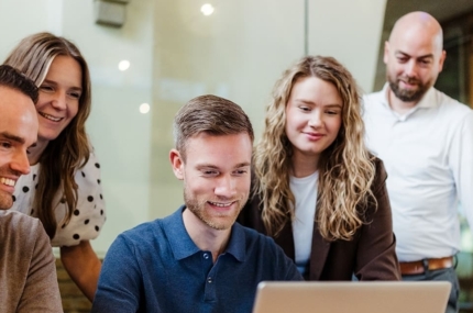Een team van vijf mensen in een vergaderruimte kijkt naar een laptop. Ze zijn gefocust op de gegevens op het scherm, met een ontspannen en gemotiveerde sfeer.