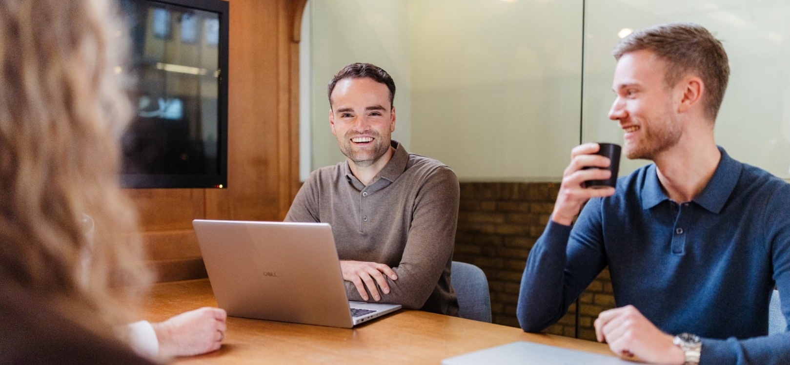 Drie collega’s zitten aan een vergadertafel met laptops en koffie en lachen tijdens een informeel overleg.