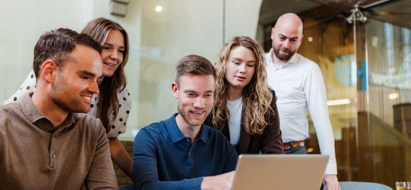Een team van vijf mensen in een vergaderruimte kijkt naar een laptop. Ze zijn gefocust op de gegevens op het scherm, met een ontspannen en gemotiveerde sfeer.