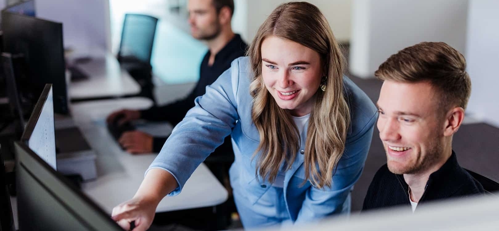 Een vrouw wijst enthousiast naar het scherm van de computer terwijl ze met een collega samenwerkt. Beide lachen terwijl ze naar het scherm kijken. Op de achtergrond is een andere collega bezig met zijn eigen werk.