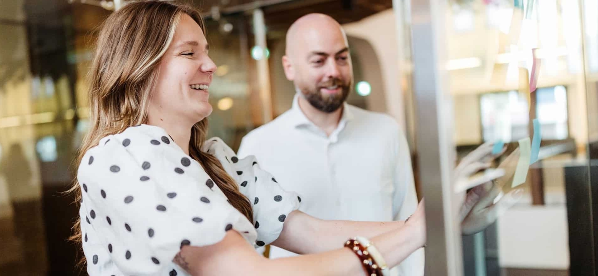 Een vrouw in een wit met zwart gestippeld shirt en een man in een wit overhemd staan samen in een vergaderruimte. De vrouw lacht terwijl ze een object aanraakt.