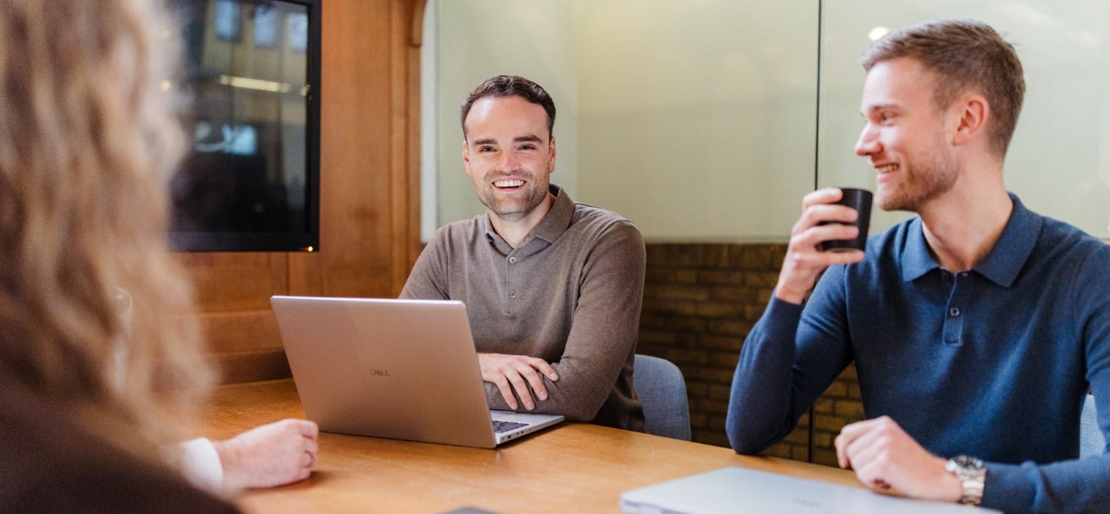 Drie collega’s zitten aan een vergadertafel met laptops en koffie en lachen tijdens een informeel overleg.