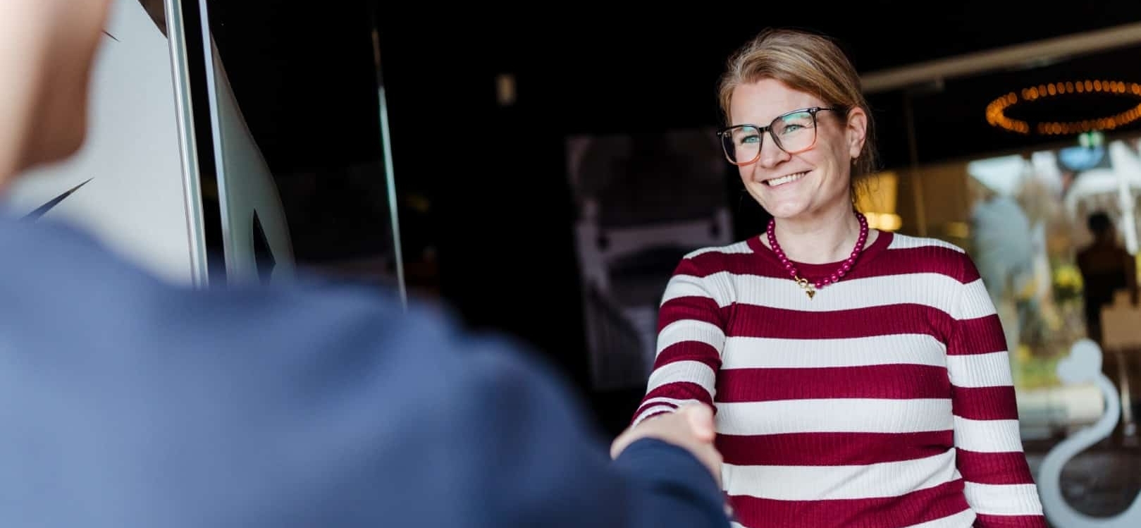 Vrouw met bril en rood-wit gestreepte trui geeft een hand bij een ontvangstbalie.