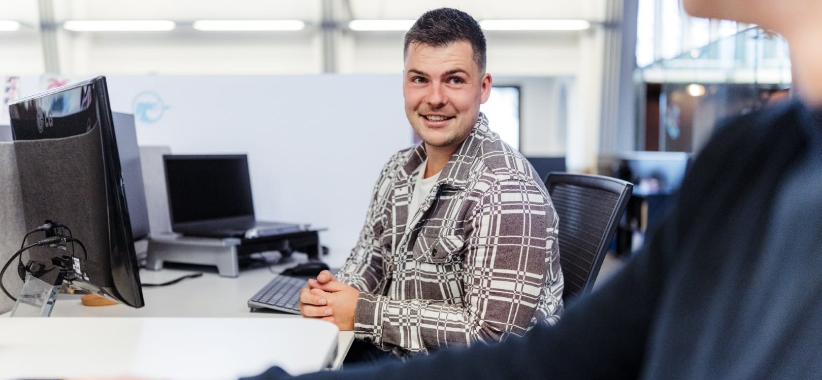 Een medewerker zit aan zijn bureau en kijkt glimlachend naar een collega in een moderne kantoorruimte