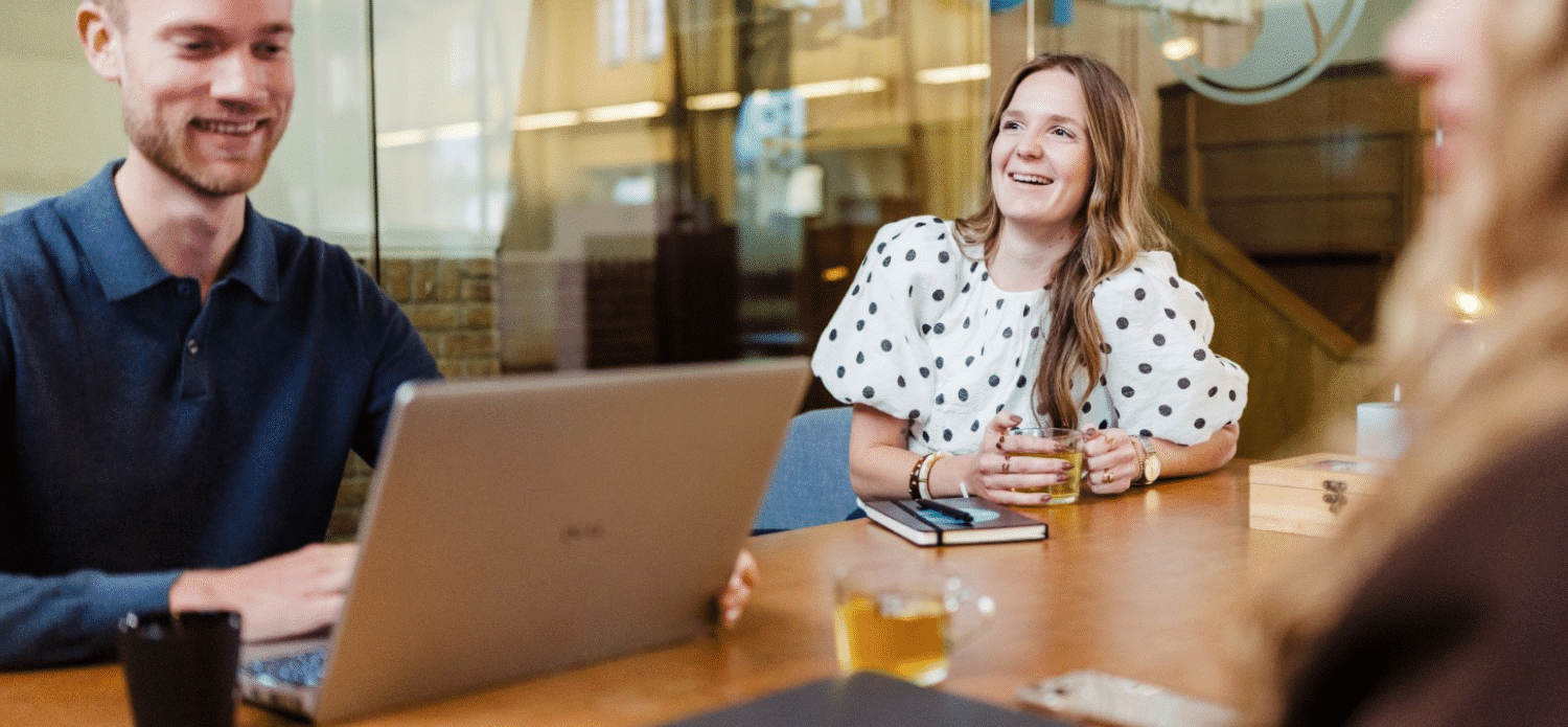 Twee collega’s zitten aan een vergadertafel met laptop, notitieboek en kopjes thee tijdens een overleg.