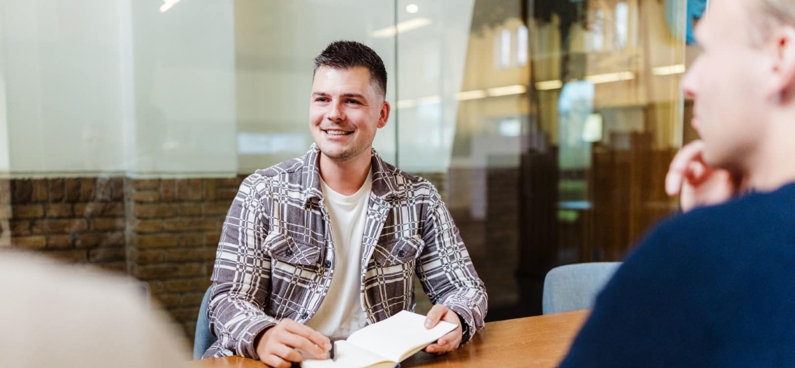 Een man zit aan een vergadertafel met een notitieboek en glimlacht terwijl hij in gesprek is met collega’s.
