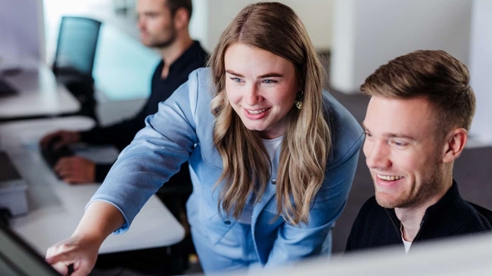Een vrouw wijst enthousiast naar het scherm van de computer terwijl ze met een collega samenwerkt. Beide lachen terwijl ze naar het scherm kijken. Op de achtergrond is een andere collega bezig met zijn eigen werk.
