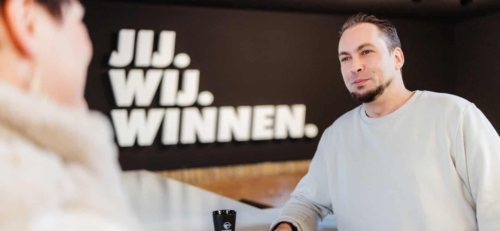 Mannelijke collega staat bij een balie op kantoor met op de achtergrond de tekst ‘Jij. Wij. Winnen.’