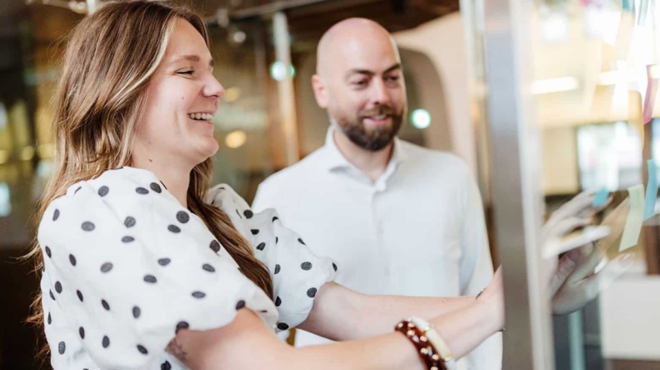 Een vrouw in een wit met zwart gestippeld shirt en een man in een wit overhemd staan samen in een vergaderruimte. De vrouw lacht terwijl ze een object aanraakt.