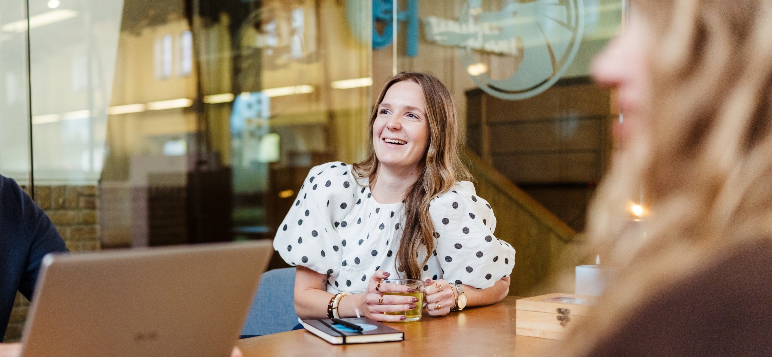 Drie collega’s zitten rond een tafel met een laptop tijdens een overleg. Ze lachen en lijken een informeel gesprek te voeren.