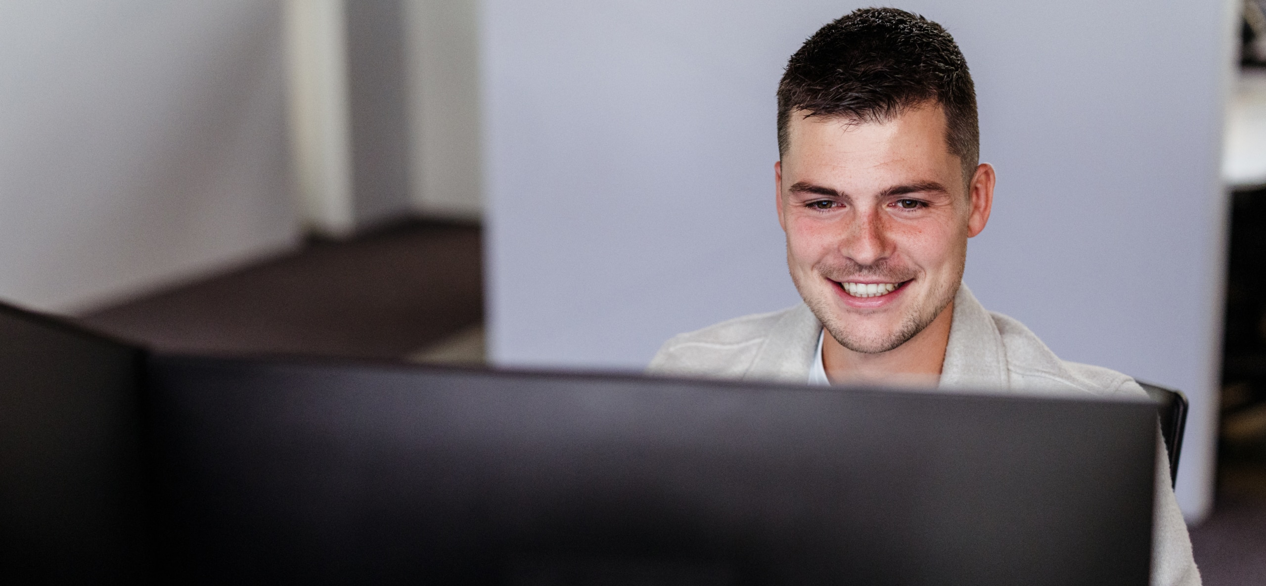 Een medewerker glimlacht terwijl hij achter een groot computerscherm werkt in een kantooromgeving.